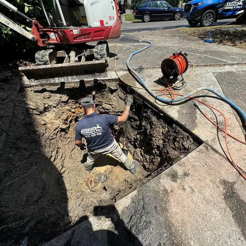 Antons plumber working on exposed drainage beside an excavator and hose reel